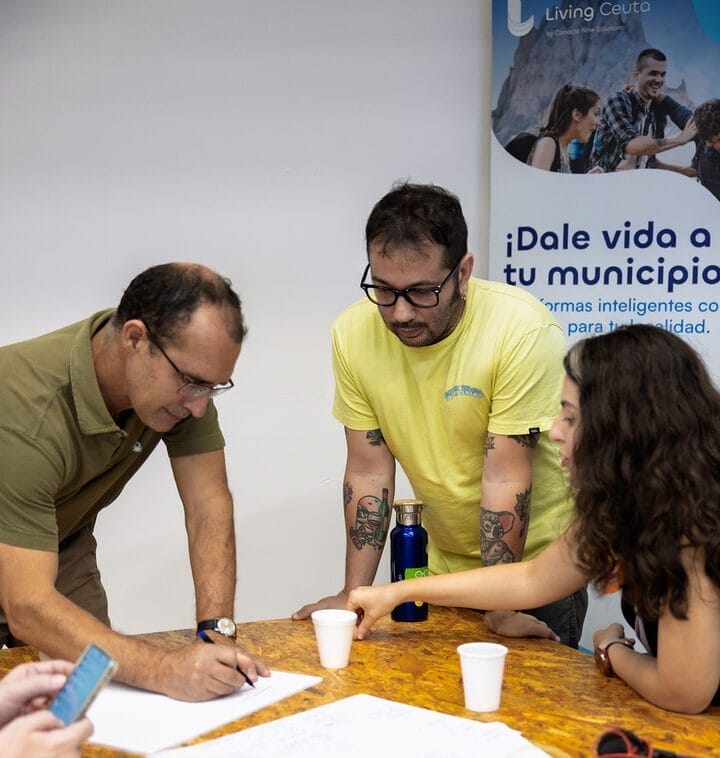 Plano medio de tres personas trabajando en una mesa de El Ángulo, Ceuta, escribiendo en papel.