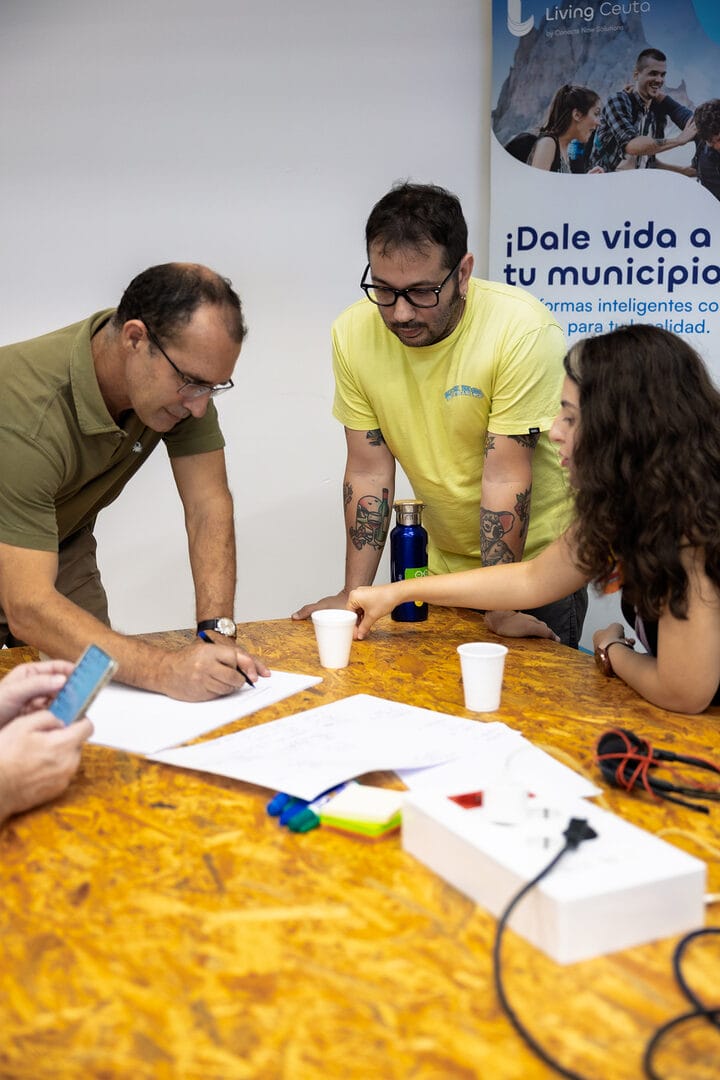 Plano medio de tres personas trabajando en una mesa de El Ángulo, Ceuta, escribiendo en papel.